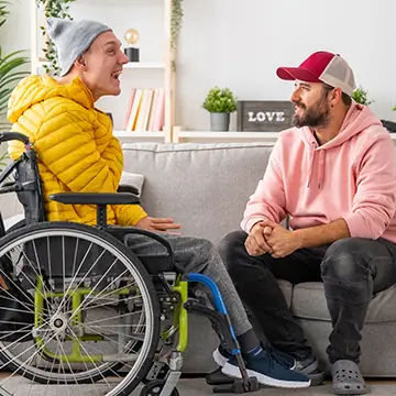 A man in a wheelchair, talking with a man sitting on a couch in a home lounge setting