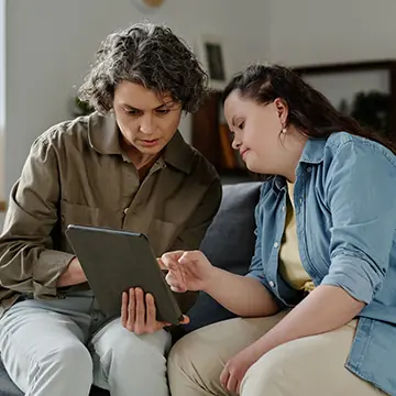 Two women sitting on a couch in a home setting, looking at a mobile tablet device