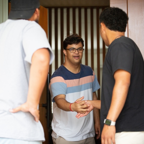A man in his early twenties greeting two people at the front door of his home.