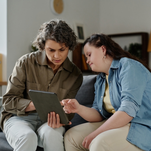 Two women sitting on a couch in a home setting, looking at a mobile tablet device