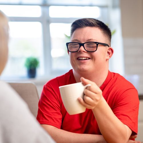 A man in his early twenties, holding a mug, talking with a woman across a table, in a home setting