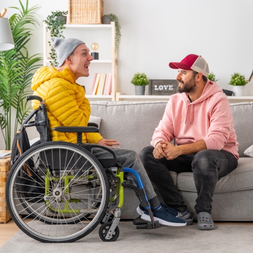 A man in a wheelchair, talking with a man sitting on a couch in a home lounge setting.